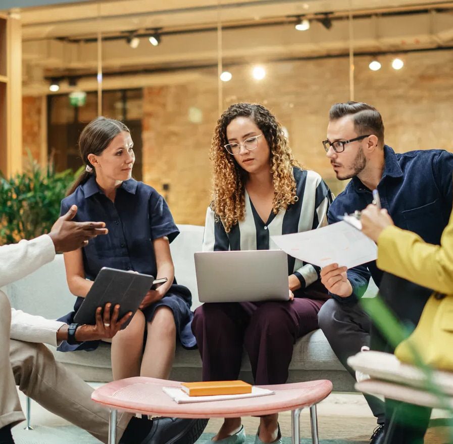 A diverse team of five colleagues meeting in a bright office lounge with laptop, tablet, and documents